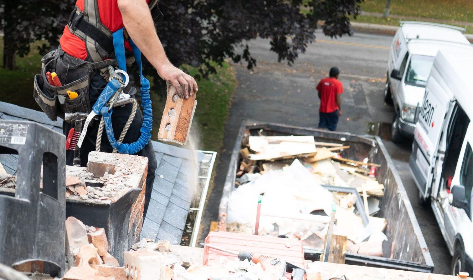 Worker in safety harness removes bricks from roof into dumpster below.